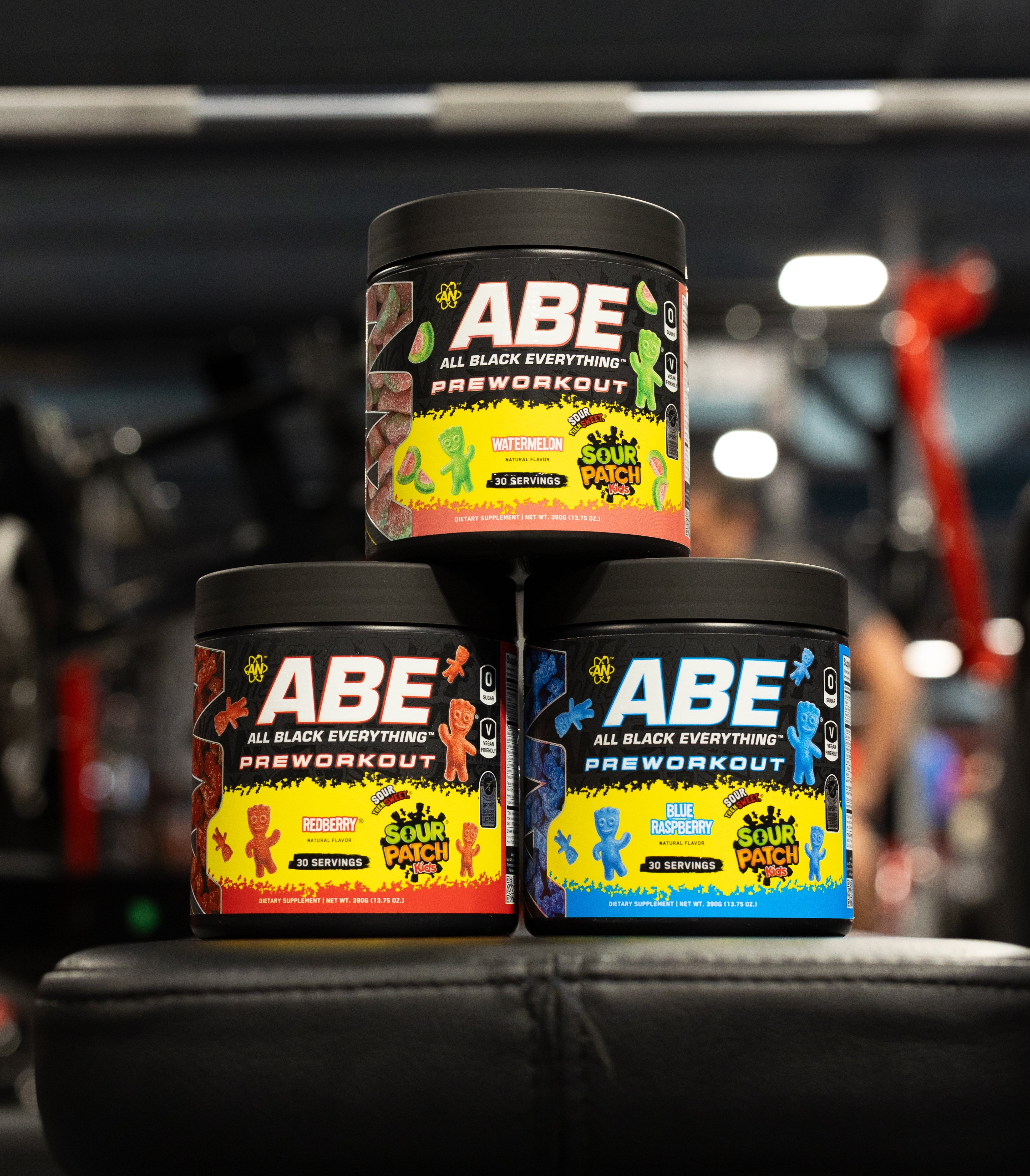 Three tubs of ABE All Black Everything Preworkout stacked on a black bench in a gym setting, with one tub on top and two on the bottom. The labels feature colorful gummy bear graphics in red, yellow, green, and blue.