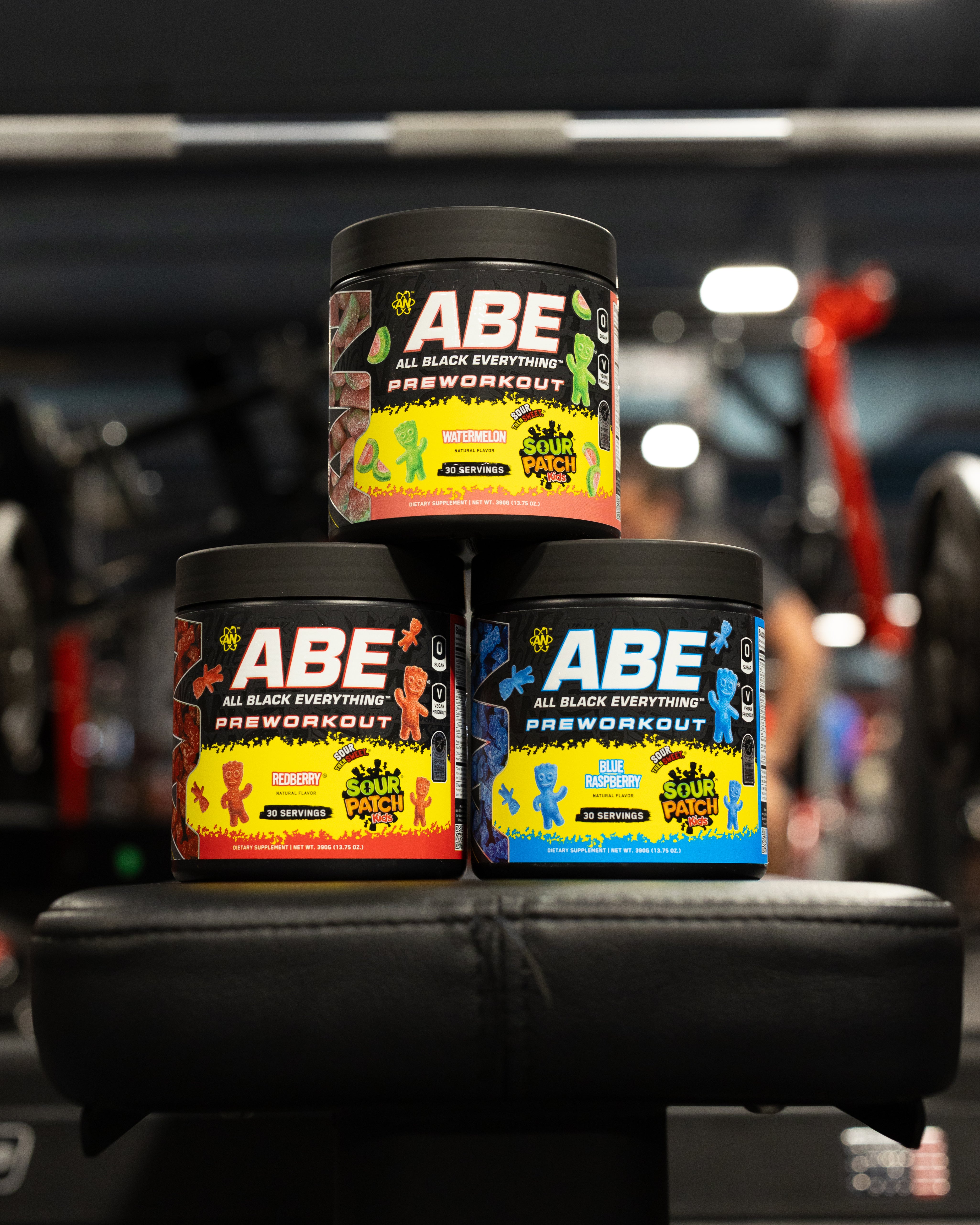 Three tubs of ABE All Black Everything Preworkout stacked on a black bench in a gym setting, with one tub on top and two on the bottom. The labels feature colorful gummy bear graphics in red, yellow, green, and blue.